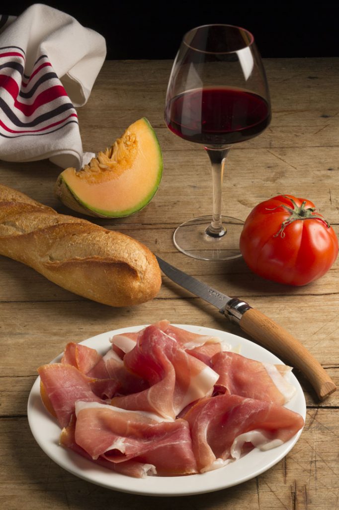 sliced bayonne ham on a white plate surrounded by bread, fruit, and wine
