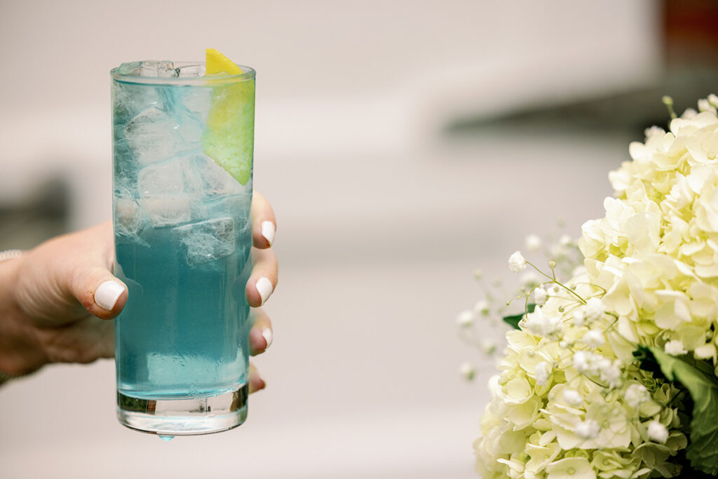 hand holding Something Blue cocktail with a slice of lemon in it near some white flowers