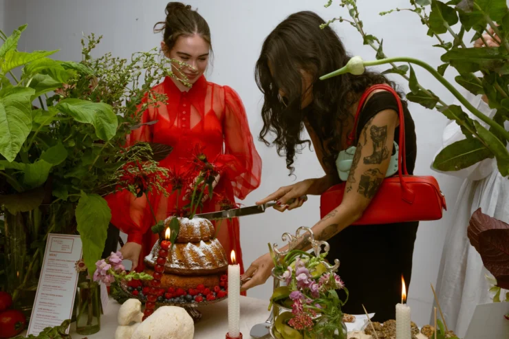 A woman with dark hair cuts into the tiered olive olive bundt cake while another woman in a red dress on the left looks on